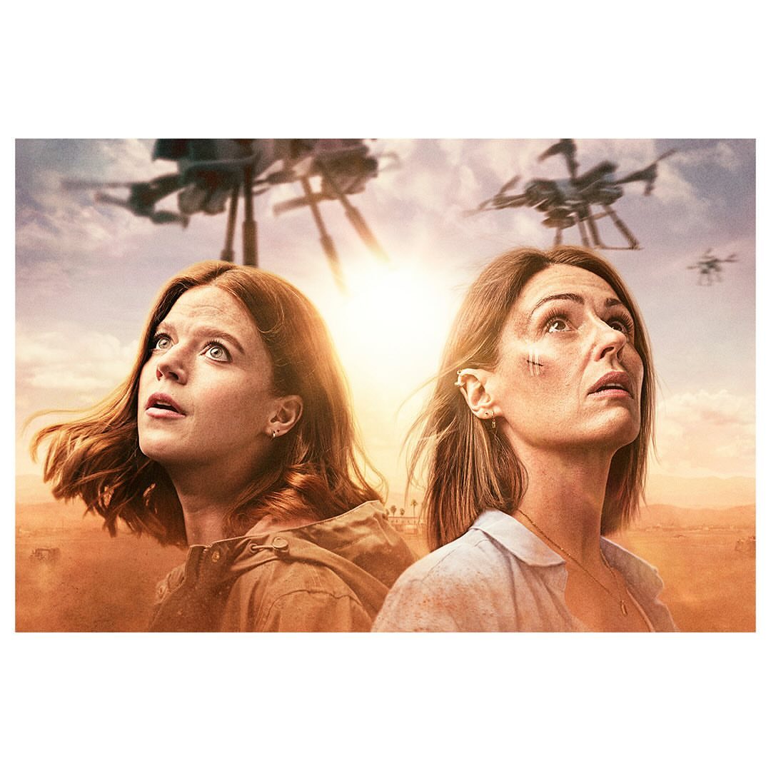 Two women gaze upward at futuristic drones in a sunlit, arid landscape, creating a scene of suspense or technological drama.