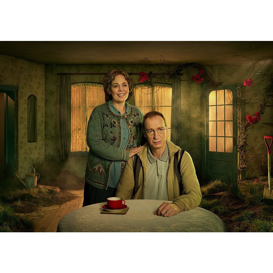 Indoor portrait of a couple at a round table with a red cup; the man is seated, the woman stands with a hand on his shoulder.