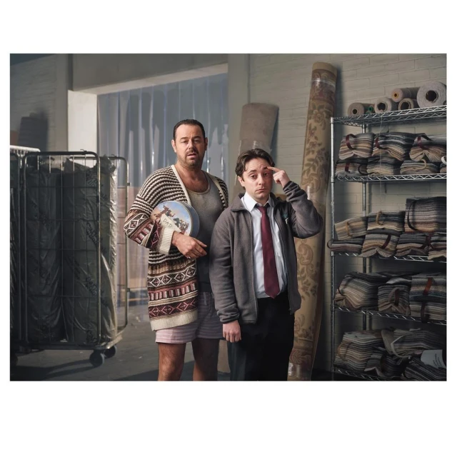 Two men stand indoors with shelves of rolled carpets. One man wears a cardigan and boxer shorts, holding a decorative plate.