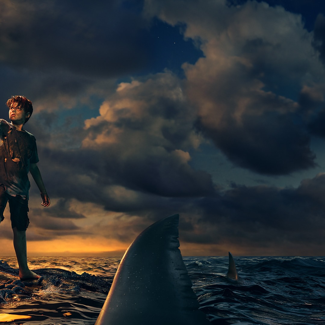 A boy is stranded on a rock surrounded by ocean waters with a shark fin visible. Dark, cloudy sky with a sliver of light on the horizon.