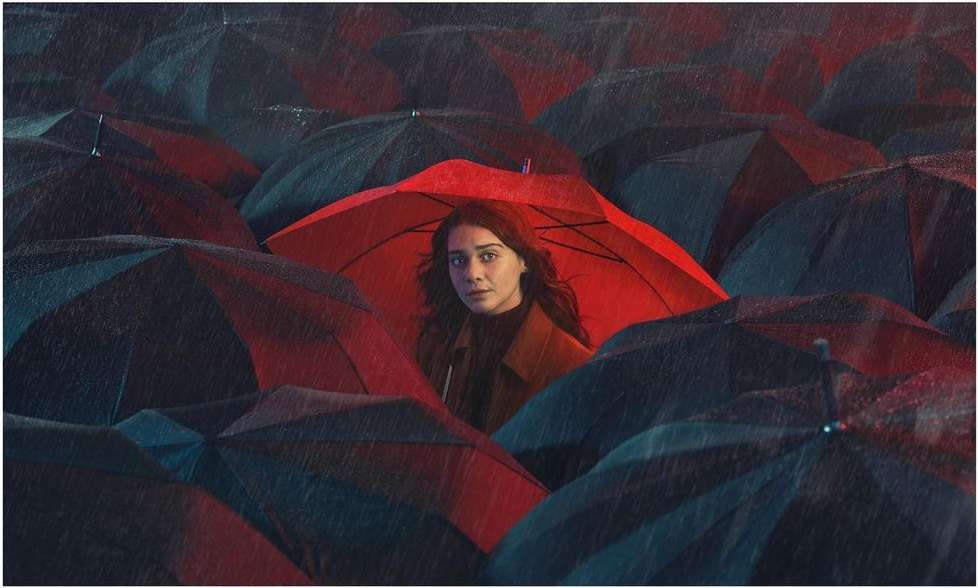 Young woman framed by a vivid red umbrella stands out amidst a sea of dark umbrellas, creating a striking contrast in an outdoor setting.
