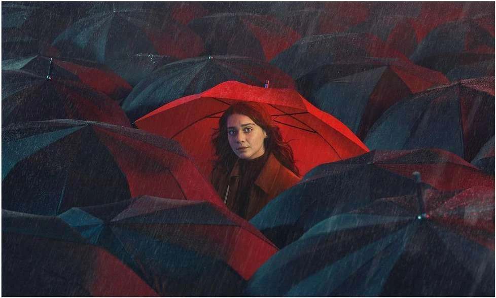 Young woman framed by a vivid red umbrella stands out amidst a sea of dark umbrellas, creating a striking contrast in an outdoor setting.