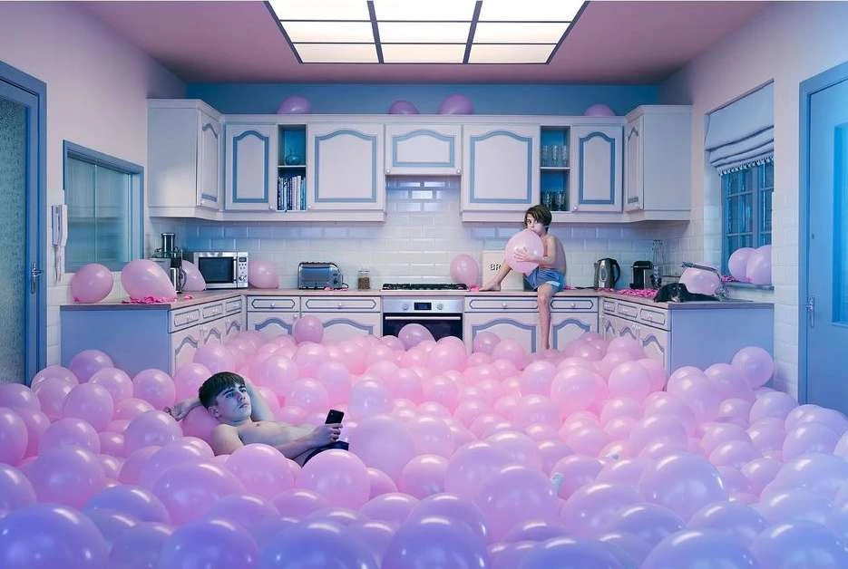 Indoor scene features two people amidst a sea of pink balloons in a kitchen with white cabinets and pastel pink and blue color scheme.