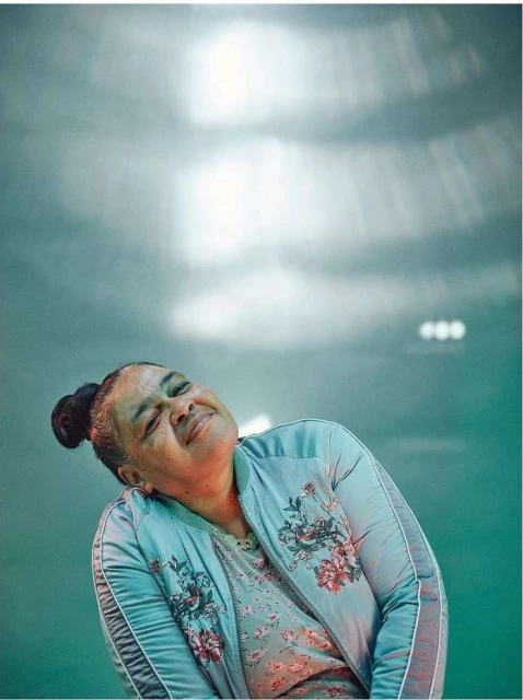 Smiling woman with bun hairstyle wearing a floral bomber jacket. Upward angle, studio lighting, soft aqua and neutral colors dominate.