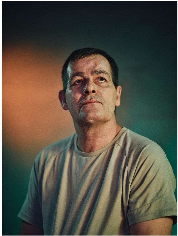 Portrait of a mature man wearing a neutral t-shirt looking upward against a dark textured background with ambient lighting.