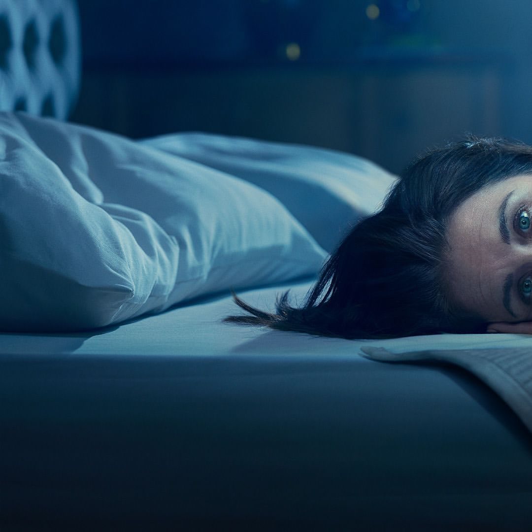 Adult woman lying in bed with blue sheets and pillow, looking up with wide eyes in a dark room, illuminated by blue lighting.