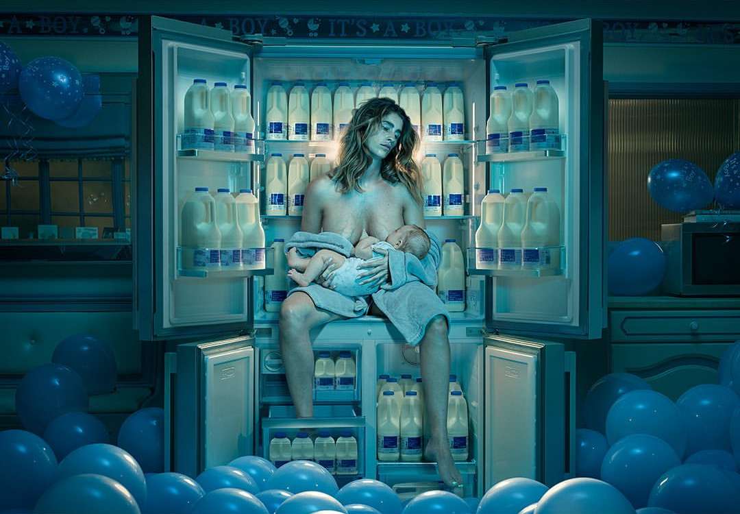 Woman breastfeeds baby sitting in front of open refrigerator stocked with milk, blue balloons in kitchen; indoor shot.