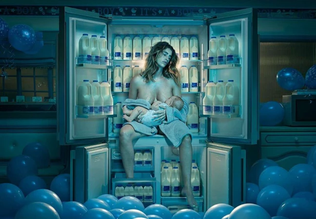 Woman breastfeeds baby sitting in front of open refrigerator stocked with milk, blue balloons in kitchen; indoor shot.