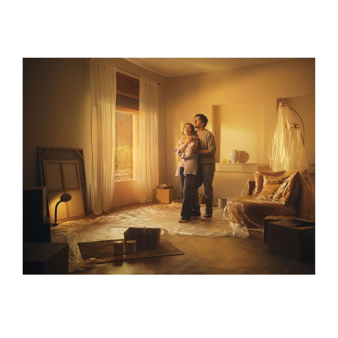 A couple in a sunlit room filled with moving supplies looks up toward the light, sharing an embrace amid a cozy interior.