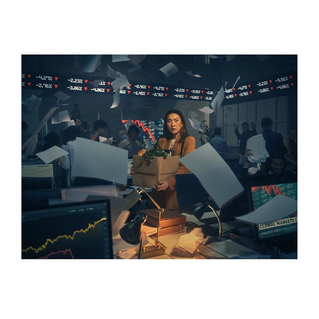 Woman walks through a chaotic office, holding a cardboard box filled with belongings, financial charts in the background.