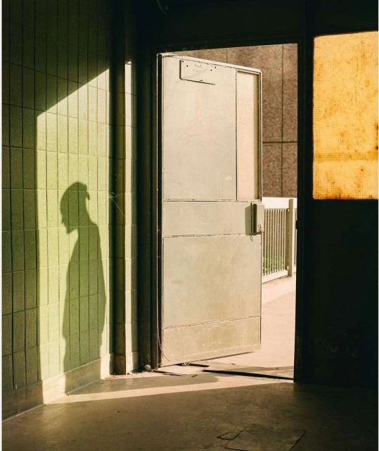 Indoor view of light illuminating tiled wall, with silhouette of a person and partially open metal door. Architectural photo.