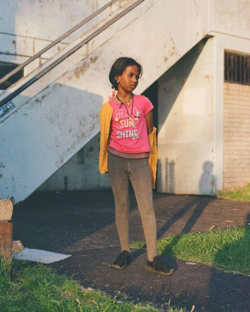 A young girl stands in front of a concrete staircase, wearing a pink shirt, grey leggings, and a yellow sweater draped over her shoulders.