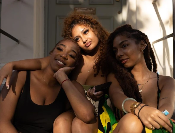 Three young women with various hairstyles sit close together on steps, bathed in sunlight, smiling and looking at the camera.