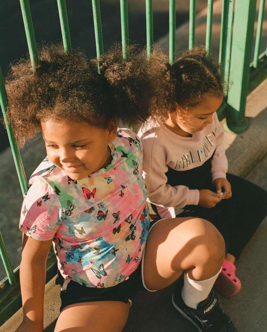 Two young girls sit side-by-side near a green fence, one in a butterfly shirt, the other in a 'Girl Power' sweatshirt, enjoying the sunlight.