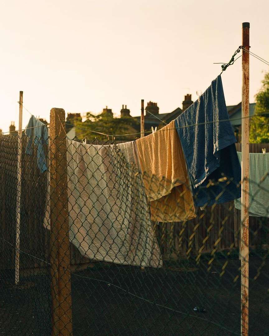 Outdoor shot of a clothesline with various clothing items drying in the sun, supported by metal posts and chain-link fence backdrop.