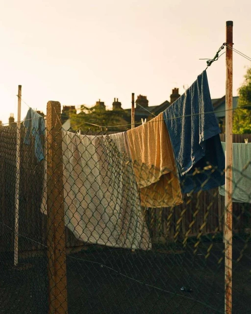 Outdoor shot of a clothesline with various clothing items drying in the sun, supported by metal posts and chain-link fence backdrop.