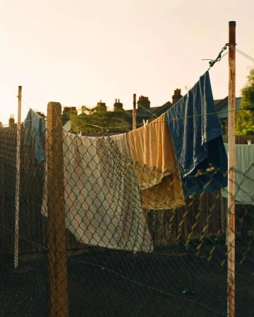 Outdoor shot of a clothesline with various clothing items drying in the sun, supported by metal posts and chain-link fence backdrop.