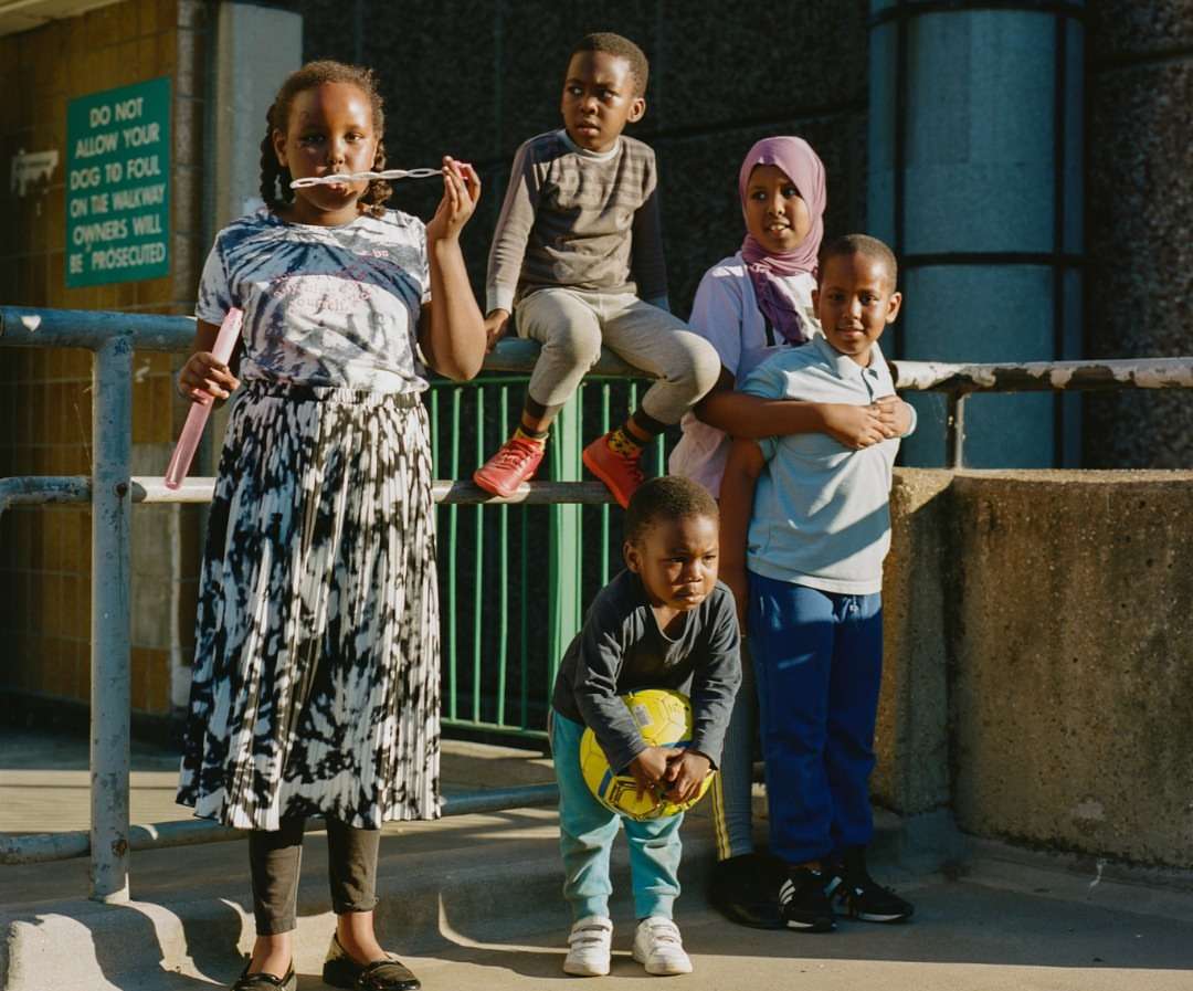Group of five children in an urban environment with one blowing bubbles and another holding a soccer ball; concrete backdrop and metal railing.