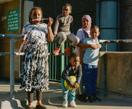 Group of five children in an urban environment with one blowing bubbles and another holding a soccer ball; concrete backdrop and metal railing.