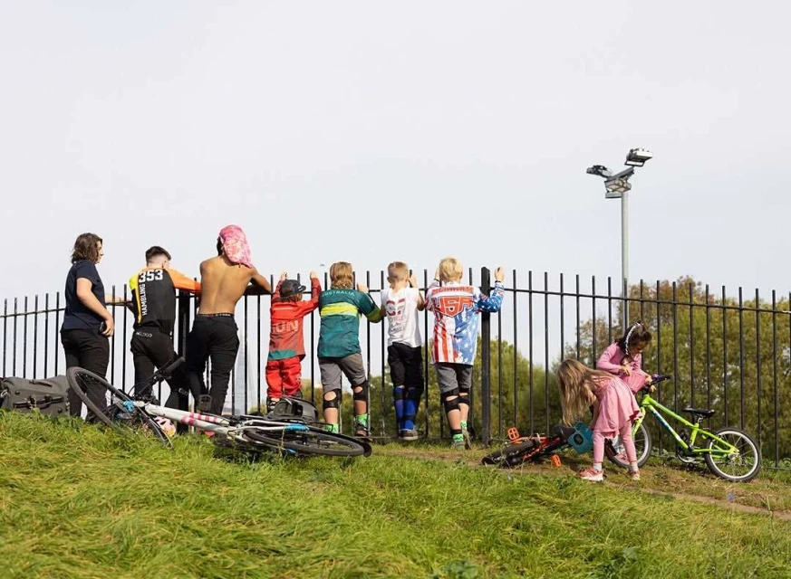 Group of children and young adults look over a fence at a grassy hill with bicycles; youth sports, active lifestyle, casual fashion.