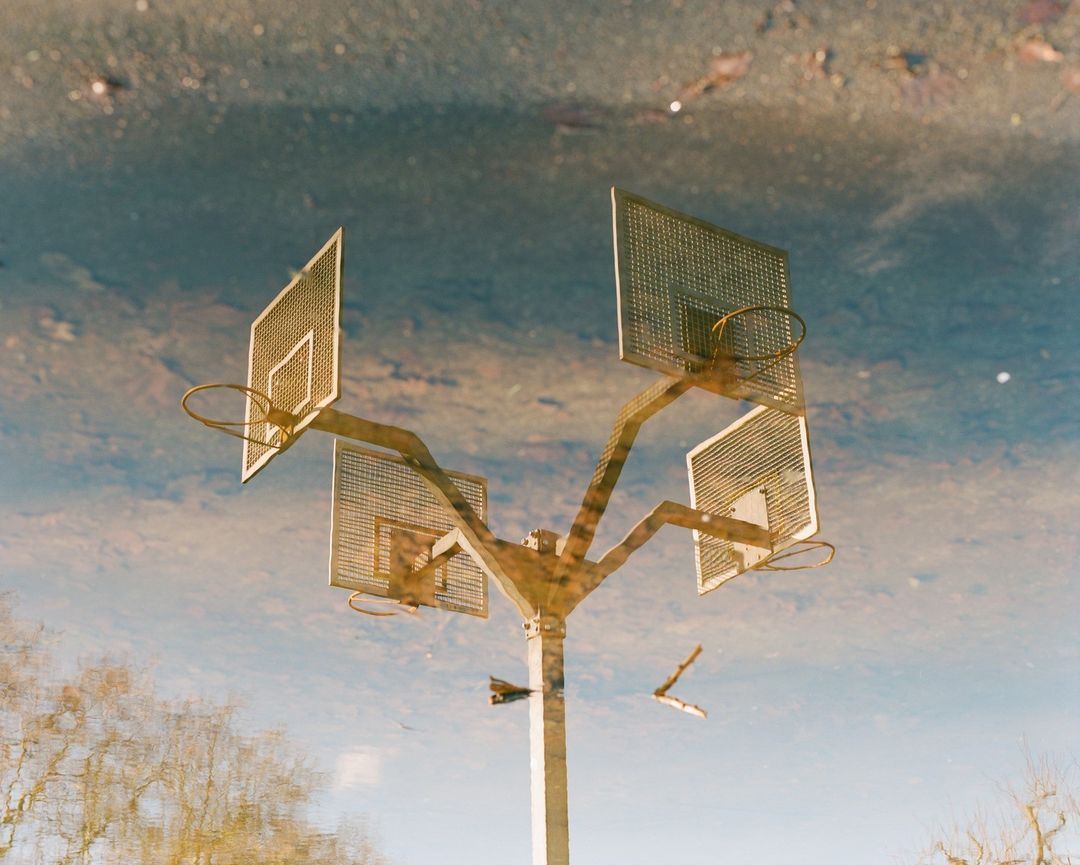 Reflection of a multi-basketball hoop structure in a puddle, capturing an outdoor scene with trees and sky visible in the background.