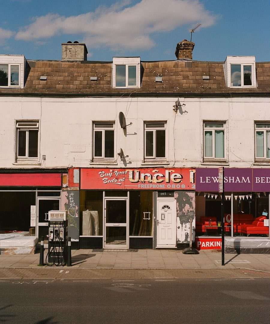 Commercial buildings line a street with a parking sign and petrol dispenser, featuring architectural facades under a sunny sky.
