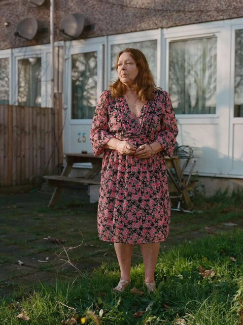 Full shot of a mature woman standing in a backyard wearing a floral dress in front of a residential house on a sunny day.