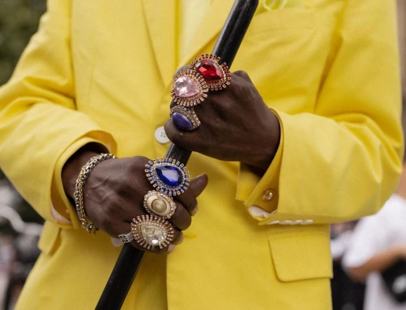 Close-up of a person wearing a yellow suit, holding a cane and displaying large rings on their fingers, showcasing jewelry fashion.