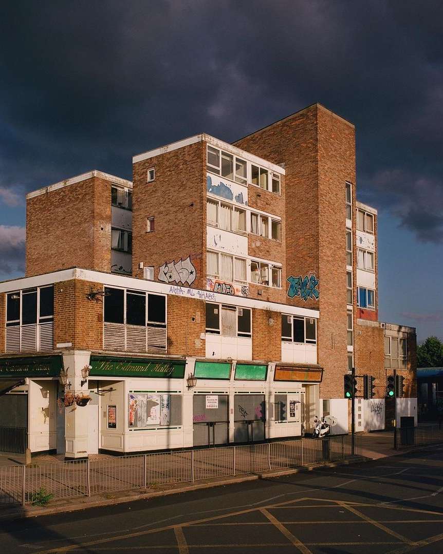 Exterior view of a multi-story brick building with a row of shops below, under a cloudy sky, in a city setting with street.