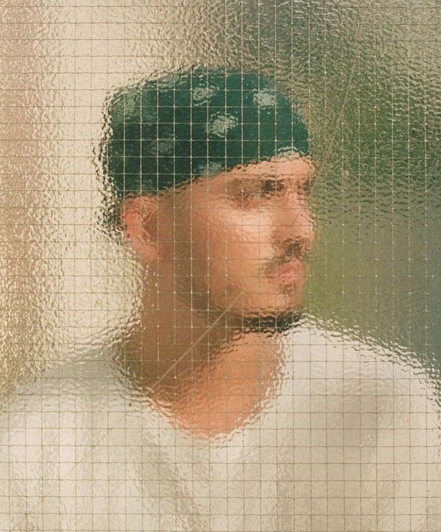 Portrait of a man with a mustache and a green bandana looking away, partially obscured by a textured glass panel. 