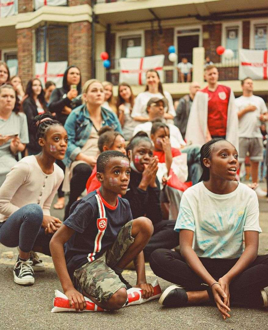 A group of diverse children and adults gathered outside watching something, with England flags decorating a building in the background.