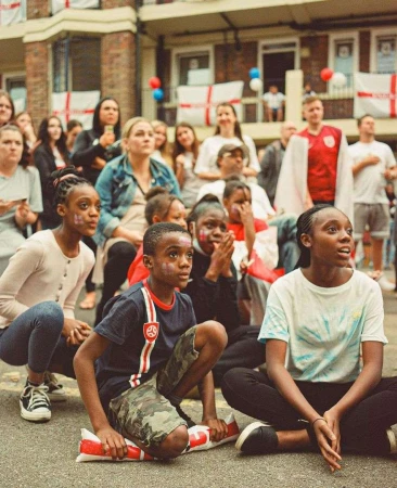 A group of diverse children and adults gathered outside watching something, with England flags decorating a building in the background.