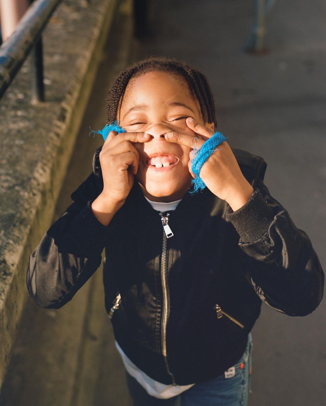 A child makes a funny face, using fingers to pull up the corners of their eyes and scrunch their nose while smiling with visible teeth.