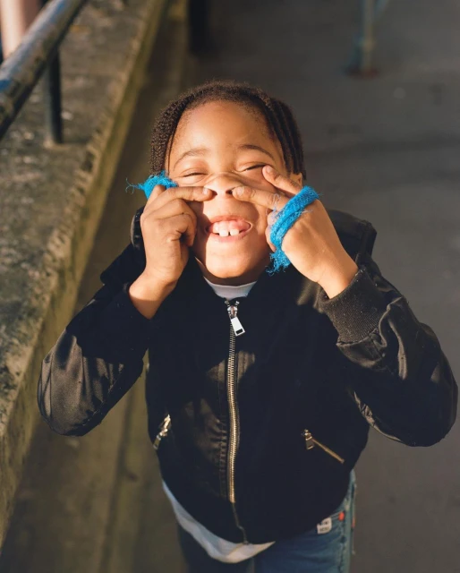 A child makes a funny face, using fingers to pull up the corners of their eyes and scrunch their nose while smiling with visible teeth.