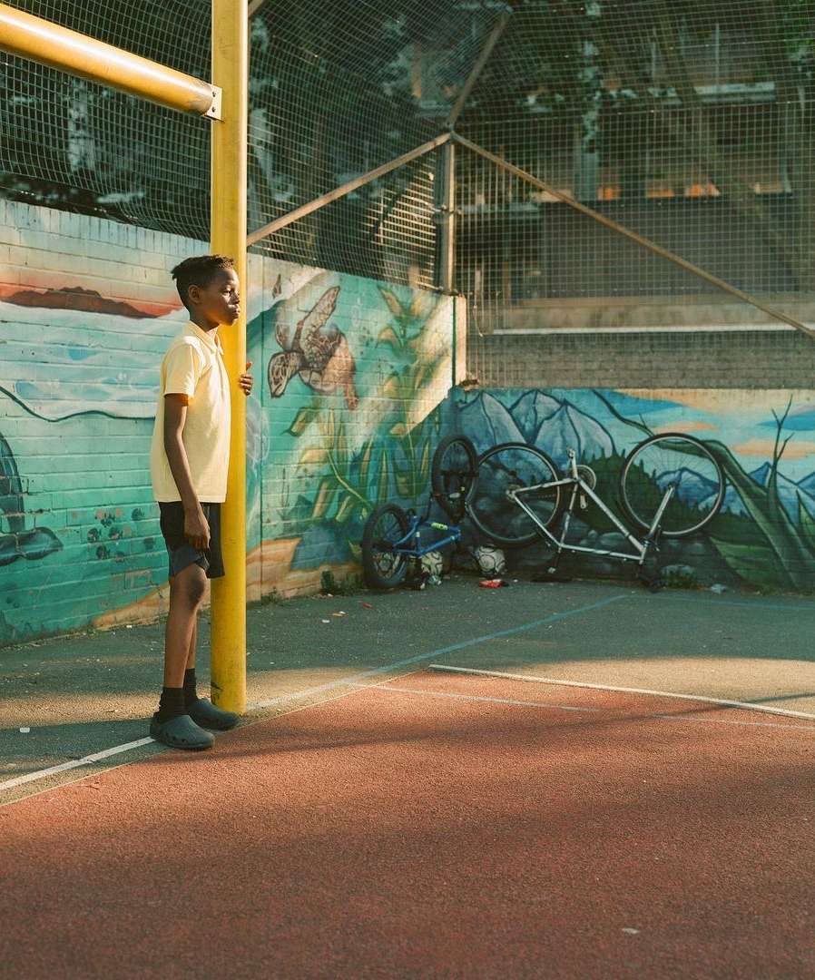Young Black boy stands next to a yellow pole on a court with a mural of nature on the wall, creating a colorful and engaging scene.