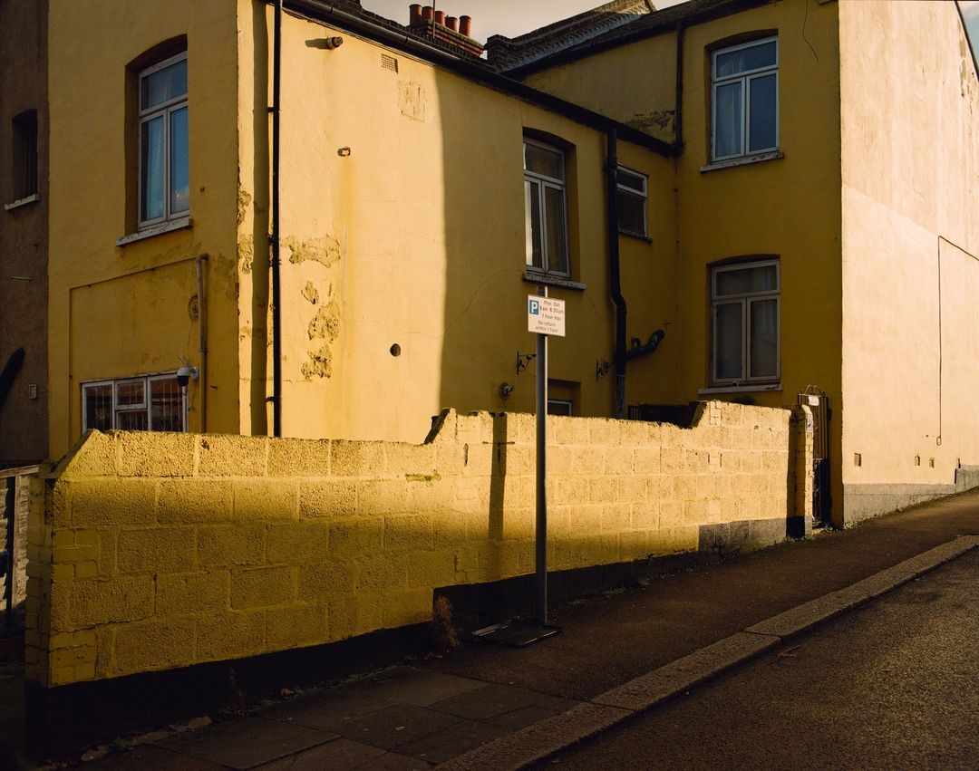 Exterior shot of yellow residential buildings and a brick wall with a parking sign on a sunny day in an urban setting.