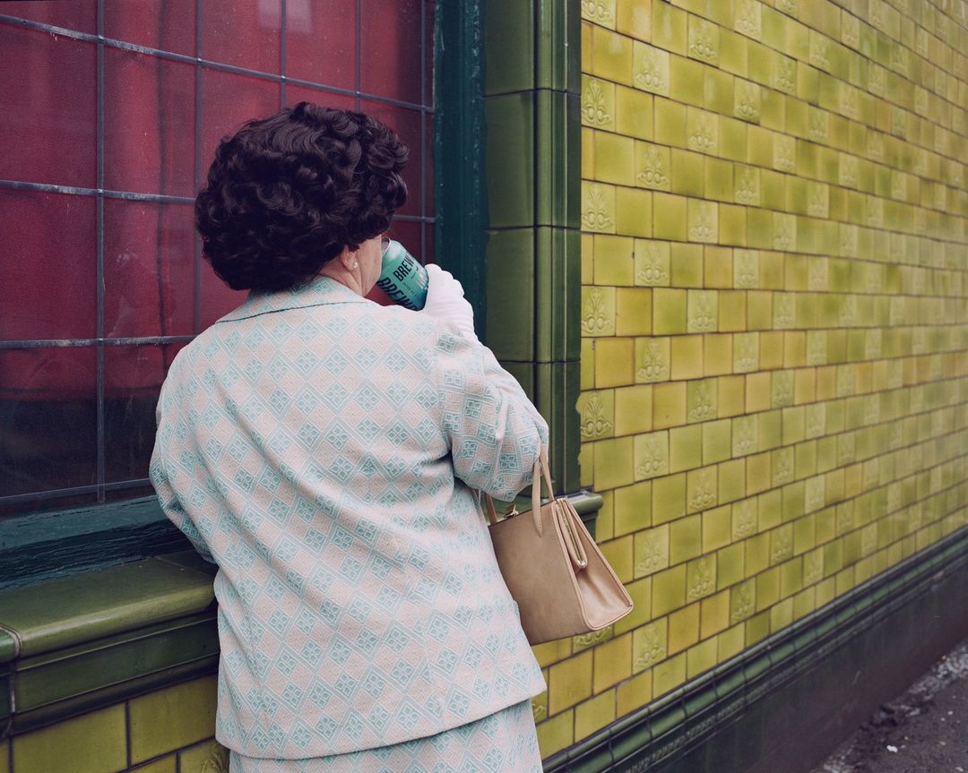 Mature woman in patterned suit drinks from can outside building with yellow tiled wall and red window. Focus on vintage street photography.