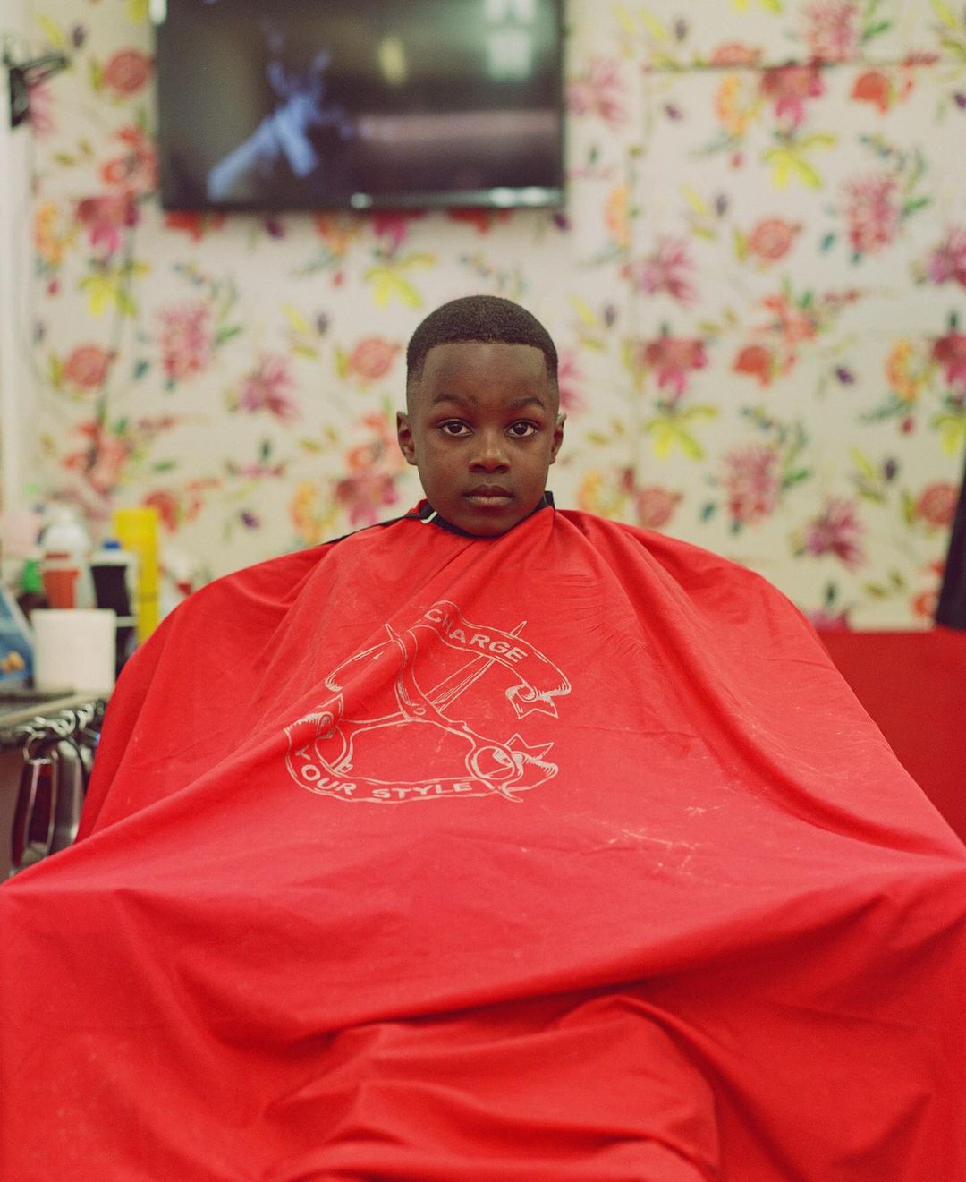 Young child wearing a red cape in a barber shop with floral wallpaper and a television screen in the background.
