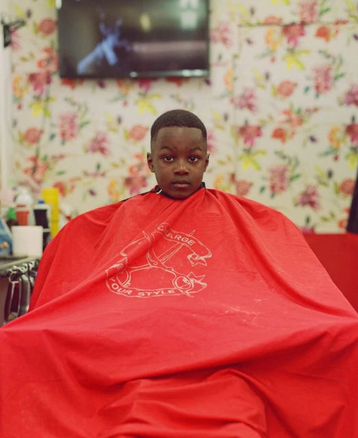 Young child wearing a red cape in a barber shop with floral wallpaper and a television screen in the background.