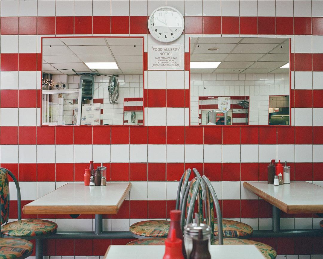 Retro diner interior with red and white striped tiled walls, mirrored openings to the kitchen, and tables with chairs.