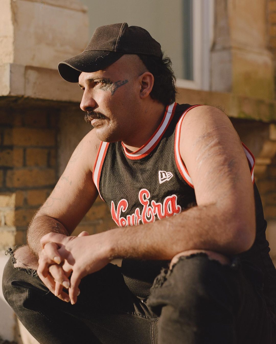 Man in Bulls jersey and cap posing outdoors, showcasing mustache, eye makeup, and unique hair. The image conveys an urban fashion style.