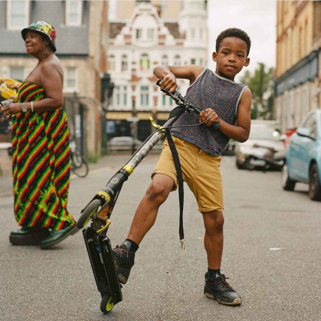 Young boy on a scooter on a city street with buildings and traffic in the background, wearing shorts, tank top, and sneakers.
