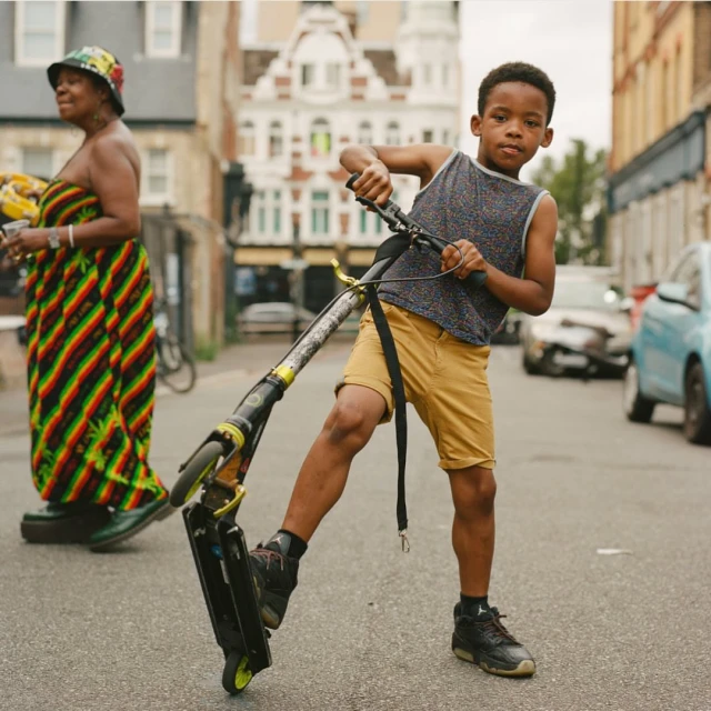 Young boy on a scooter on a city street with buildings and traffic in the background, wearing shorts, tank top, and sneakers.