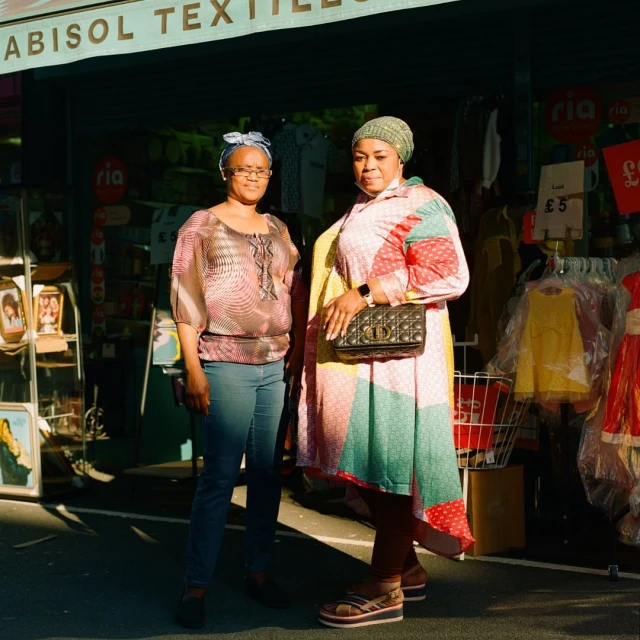 Two stylish adult women stand together outdoors next to a shopfront, wearing colorful clothing and head coverings, during daytime.