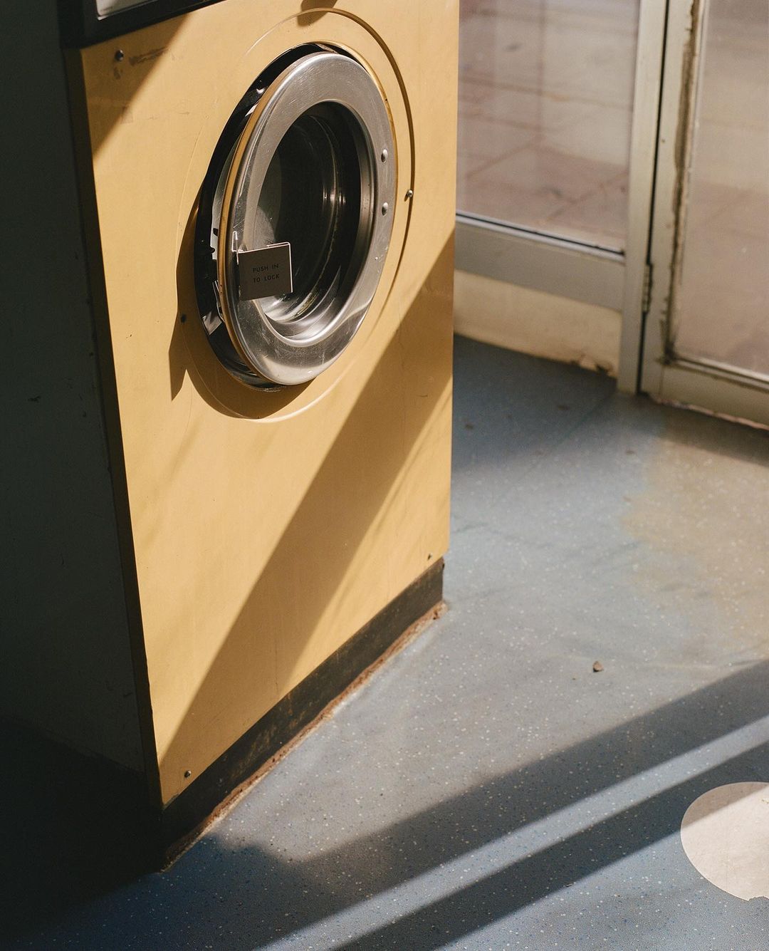 Commercial laundry machine in a laundromat bathed in natural light and shadow creating geometric patterns on the floor.