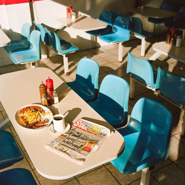 Breakfast meal of eggs, beans, and fries with coffee and newspaper on a table in a brightly lit restaurant with blue chairs.