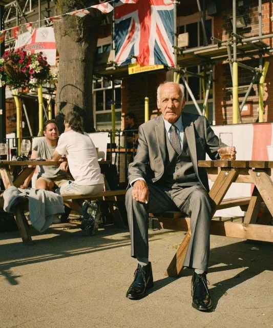 Elderly man in suit sits on a wooden bench outdoors, holding a beer; Union Jack flags hang in the background, bright daylight.