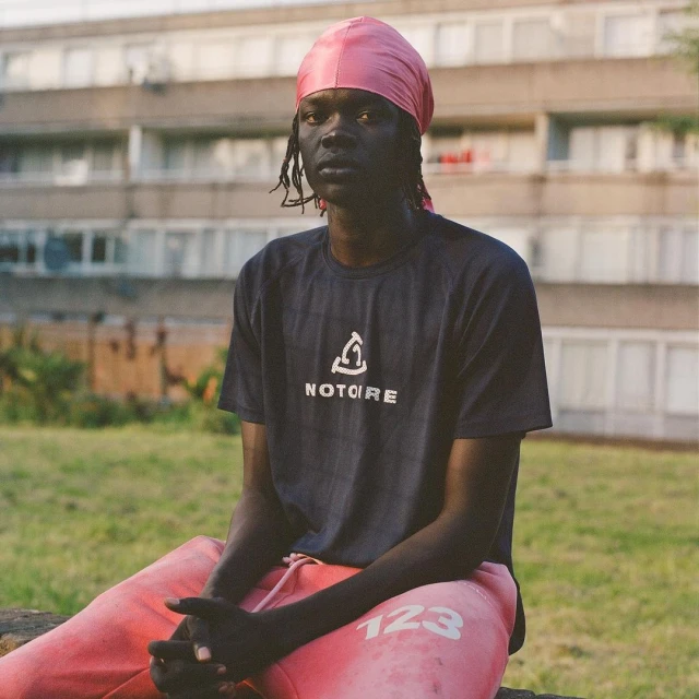 An adult Black man wearing a pink headscarf and dark t-shirt with light pink sweatpants, sitting outdoors with a blurred building background.