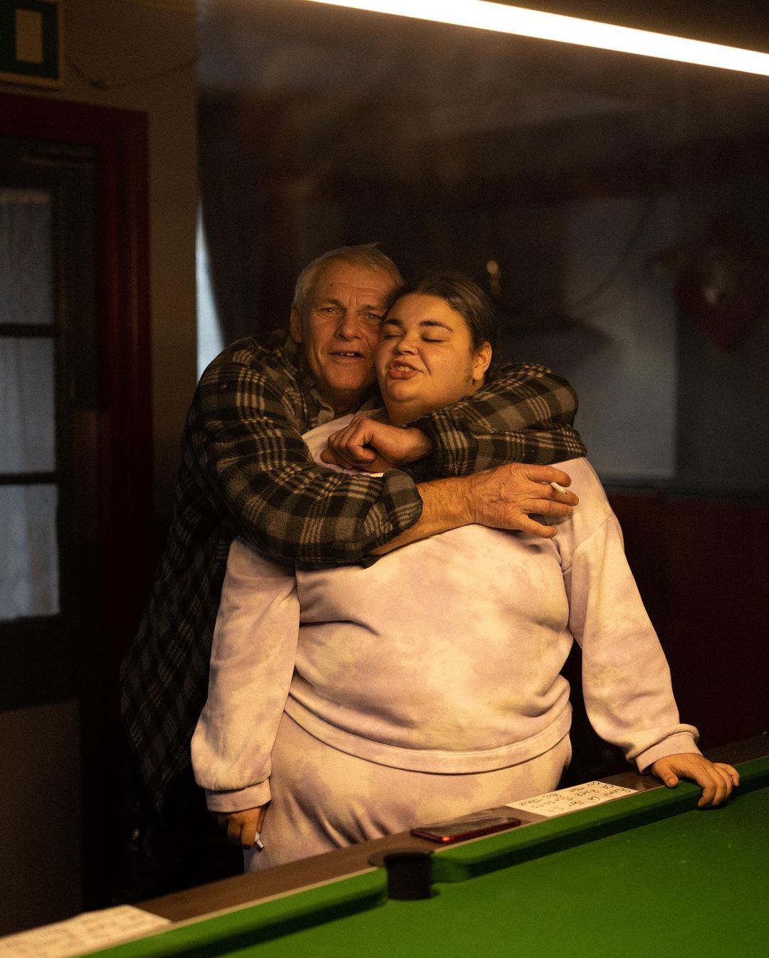 Indoor shot features an elderly man embracing an adult woman, both near a green pool table, warmly lit from above.