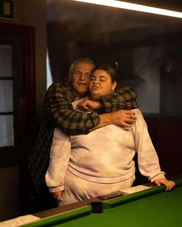 Indoor shot features an elderly man embracing an adult woman, both near a green pool table, warmly lit from above.
